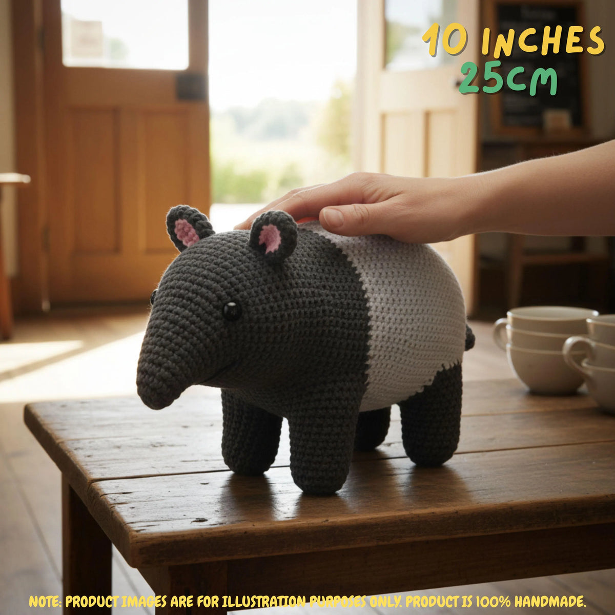 A crocheted animal pig toy with a dark gray and white body, pink inner ears, and black legs, standing on a wooden table with a human hand resting on its back. The background shows a warm, sunlit room with wooden doors and stacked ceramic cups.