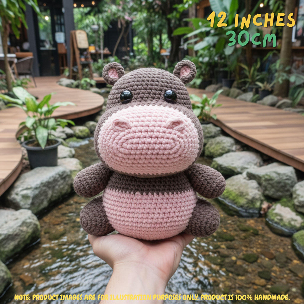 A crocheted hippo plush toy held in a person's hand in an outdoor garden setting with a stream and wooden walkways. Text overlays indicate the size as 12 inches / 30 cm, with a note at the bottom stating product images are for illustration purposes only.