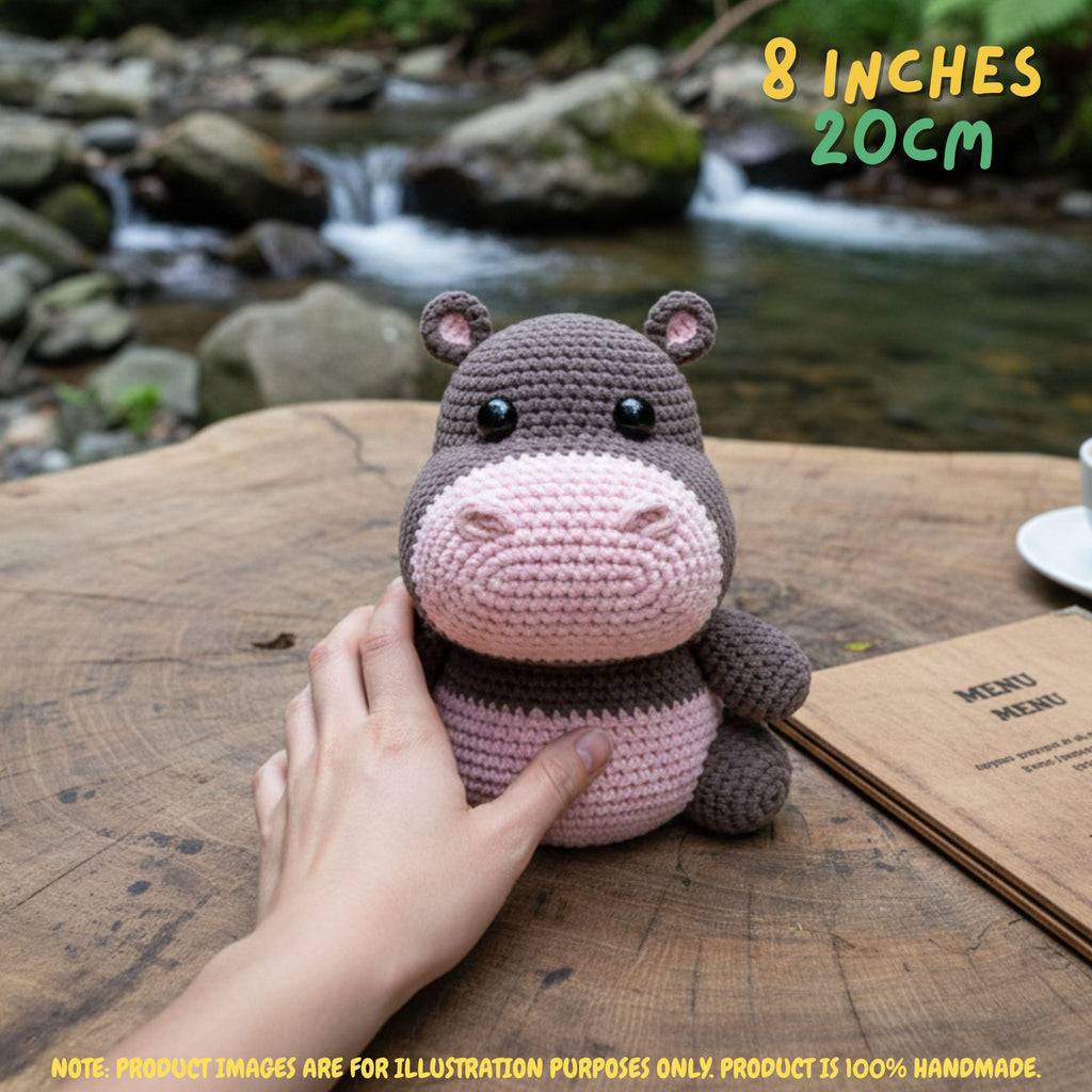 A crocheted hippo plush toy being held by a hand on a wooden table, with a blurred stream and rocks in the background. Yellow text in the top right reads '8 INCHES 20CM'.