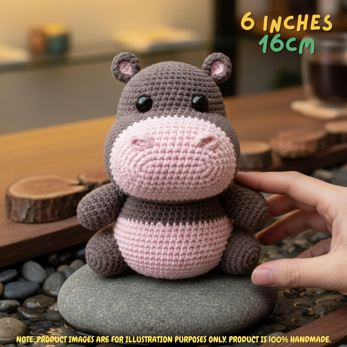 Crocheted hippo toy being held by a human hand, sitting on a stone in a styled indoor setting. Text in the top right reads '6 INCHES 16CM' and there is a note at the bottom stating product images are for illustration purposes only.