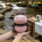 A crocheted hippo plush toy being held in two hands, with a blurred outdoor stream and wooden table in the background. Yellow text in the top right reads '10 INCHES 25CM', and a note at the bottom says product images are for illustration purposes only.