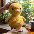 A yellow crocheted plush duck toy being held by two hands on a wooden windowsill, with a glass teapot and green garden foliage in the background. Text overlays indicate the size as 10 inches (25cm) and note that the product is 100% handmade.