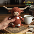 A crocheted toy longhorn bull being held by a person's hand on a wooden table, with a coffee cup, book, and coffee jar in the background. Text overlays indicate the size as 6 inches / 16 cm.