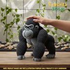 A crocheted gorilla toy is shown standing on a wooden surface with a human hand resting on its head. The background features a tiled wall with hanging green plants and decorative stones.