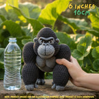 A crocheted gorilla toy is shown outdoors on a wooden surface with a hand holding it and a clear plastic bottle beside it for scale. Green leaves fill the background, and text in the top right reads '6 INCHES 16CM'.