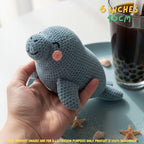 A hand holds a small crocheted seal toy in a bright indoor setting. The toy is light blue-gray with a pink cheek and simple embroidered facial features.