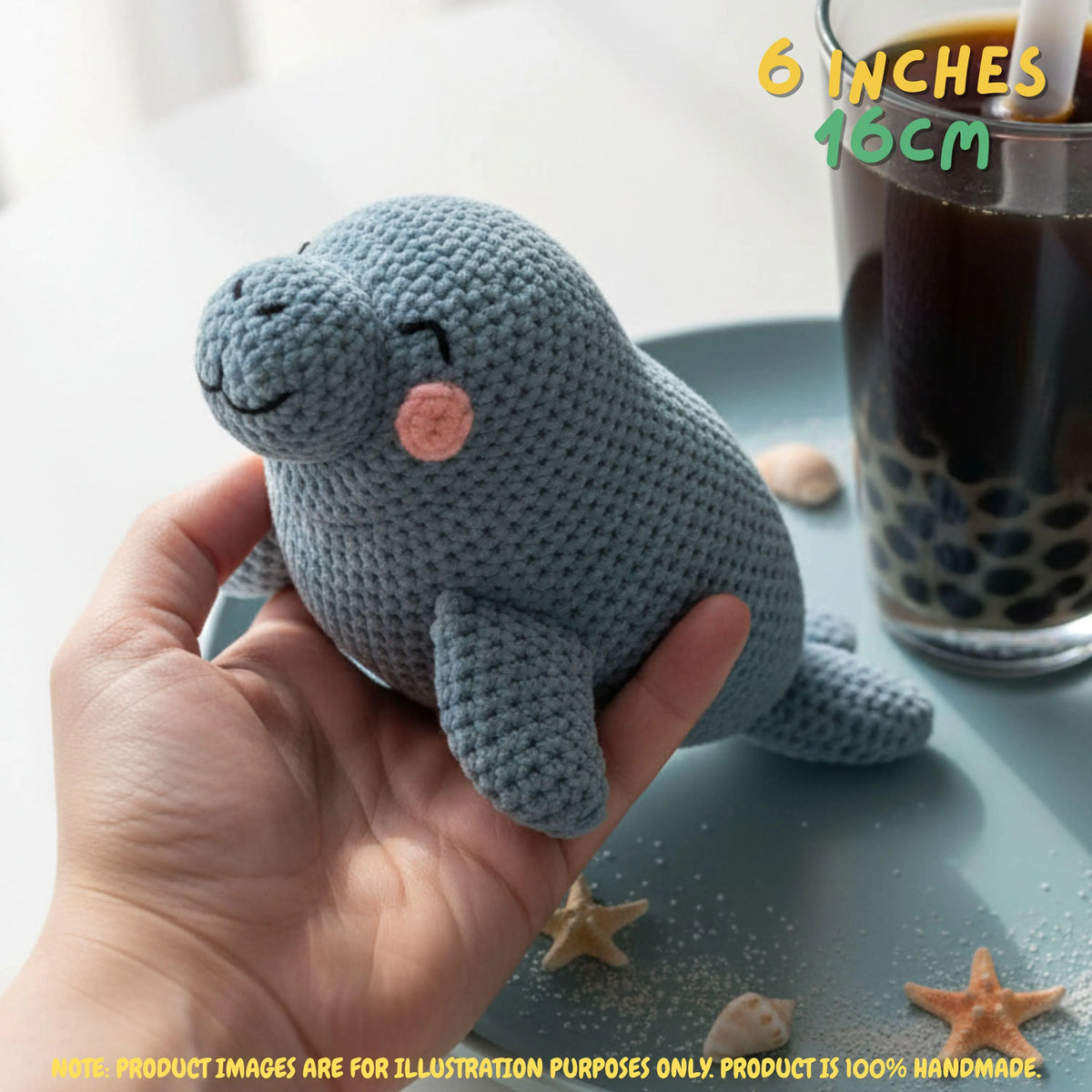 A hand holds a small crocheted seal toy in a bright indoor setting. The toy is light blue-gray with a pink cheek and simple embroidered facial features.