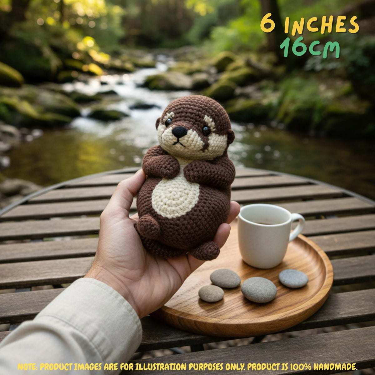 A hand holds a crocheted otter toy in front of a blurred outdoor stream scene. The otter is brown with cream-colored paws and belly.