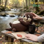 A crocheted otter plush toy is posed on a wooden bench outdoors by a stream, with a person's hands touching it. A black mug, an open book, and fallen leaves are nearby, and the background shows a blurred forest stream scene.