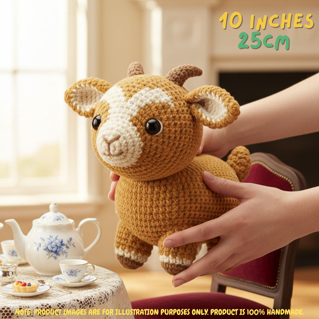 A crocheted stuffed goat toy being held by two hands in a warm indoor setting. The toy has a tan-brown body with cream and brown horns/ears and black eyes.