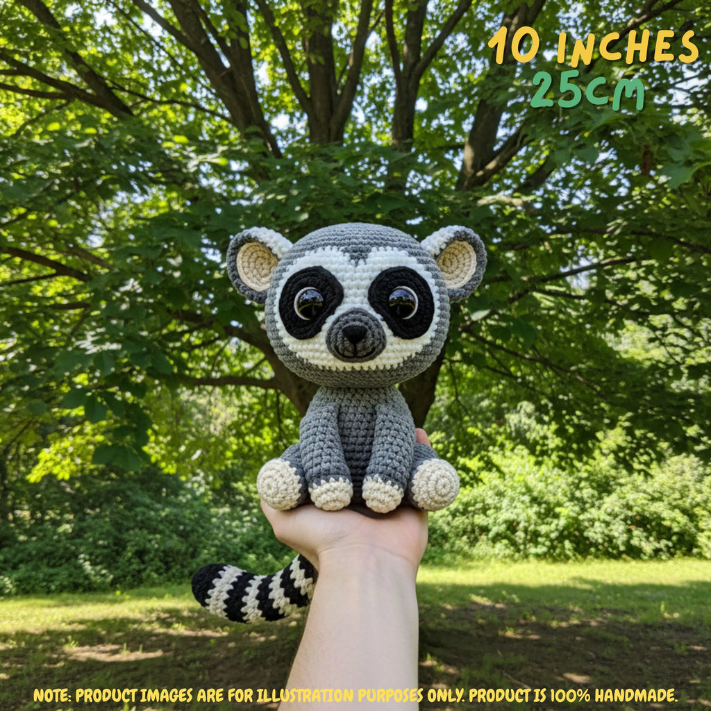 A crocheted raccoon plush toy held up by a person's hand outdoors in front of a leafy green tree. Text in the top right reads '10 INCHES 25CM', and a note at the bottom says product images are for illustration purposes only, with the product being 100% handmade.