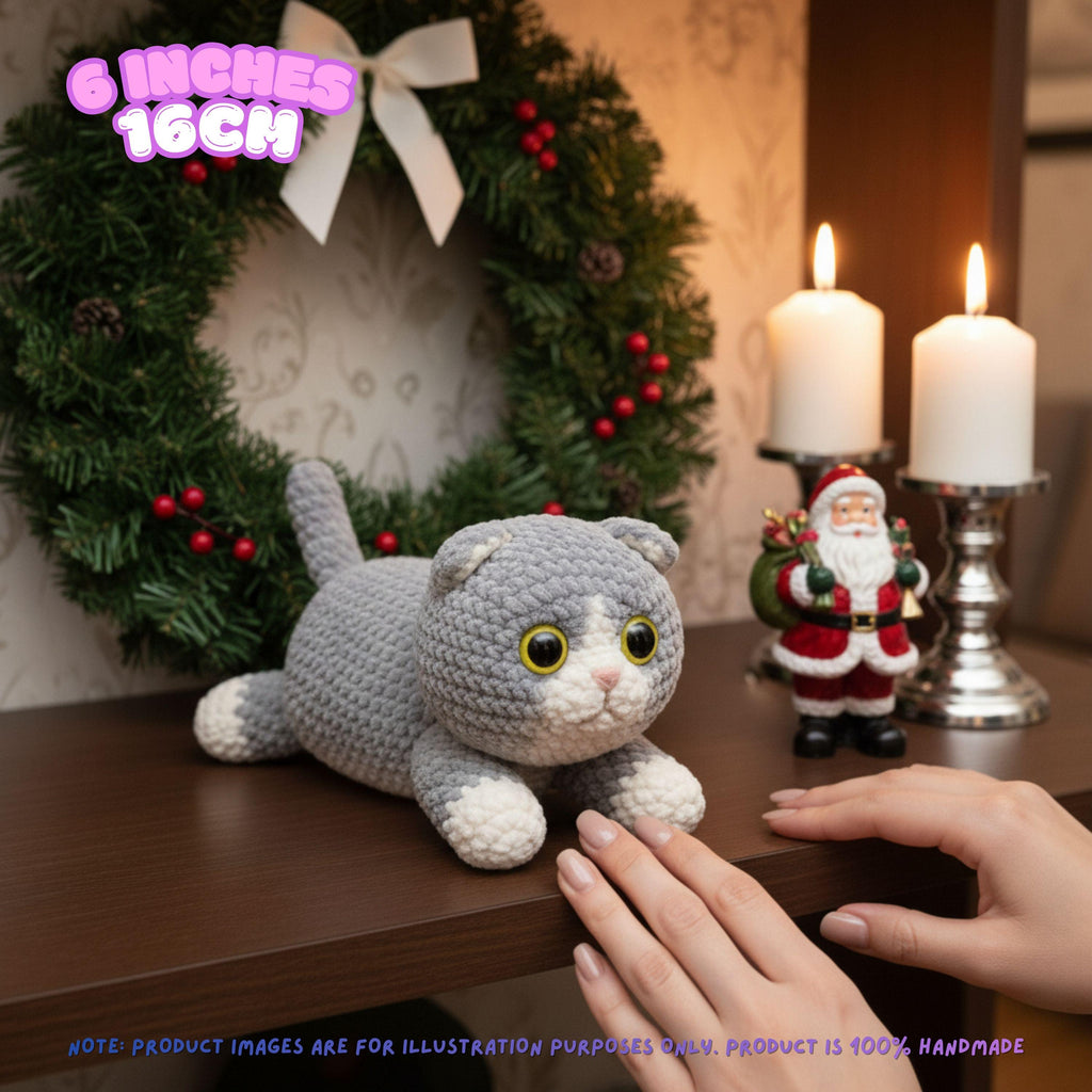 A crocheted gray and white cat toy is displayed on a wooden table in a cozy holiday setting with a green wreath, lit candles, and a small Santa figurine. A hand is reaching toward the toy, and text overlays indicate the size is 6 inches (16cm) and that the product is 100% handmade.