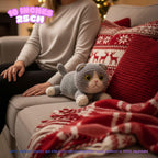 A gray and cream knitted cat plush toy rests on a light gray sofa next to a person wearing a beige sweater, with red and white holiday pillows and a blanket in the background. Text overlays indicate the size is 10 inches (25cm) and note that product images are for illustration purposes since the product is 100% handmade.