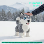 A hand-crocheted husky dog toy standing in the snow, with a human hand gently touching its head. The background shows a snowy outdoor scene with blurred trees and mountains.