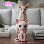 A crocheted pink cat figurine sits on a wooden table in front of a real living room background with a sofa, books, and a small plant. A real hairless cat stands behind the figurine.