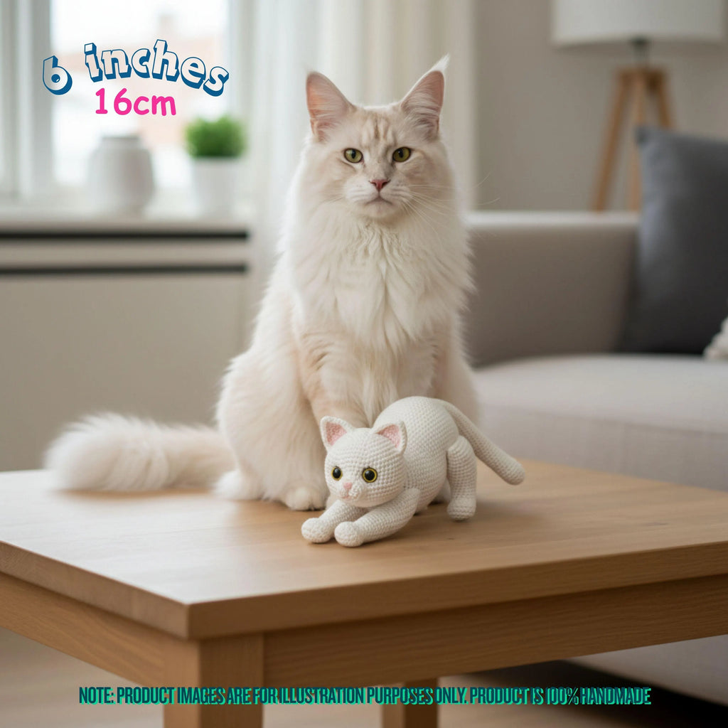 A fluffy cream-colored cat sits on a wooden table next to a small white knitted cat toy, with a blurred living room background. Text in the top left reads '6 inches 16cm'.