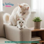 A fluffy white cat sits on a beige armchair next to a small crocheted white cat toy. The scene is indoors with a wooden side table and potted plant in the background.