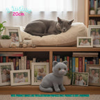 A gray crocheted cat figurine sits on a wooden shelf in front of a quilted cream pet bed where a real gray cat is sleeping. The scene includes many framed family photos, books, plants, and decorative objects, with text overlays reading '8 inches 20cm' and a note that the product image is for illustration purposes only and the product is handmade.