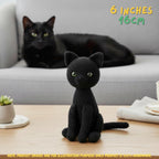 A black crocheted amigurumi cat sits on a wooden table in the foreground, with a real black cat resting on a light gray sofa in the background. Text in the top right reads '6 INCHES 16CM', and a note at the bottom says the product images are for illustration purposes only.