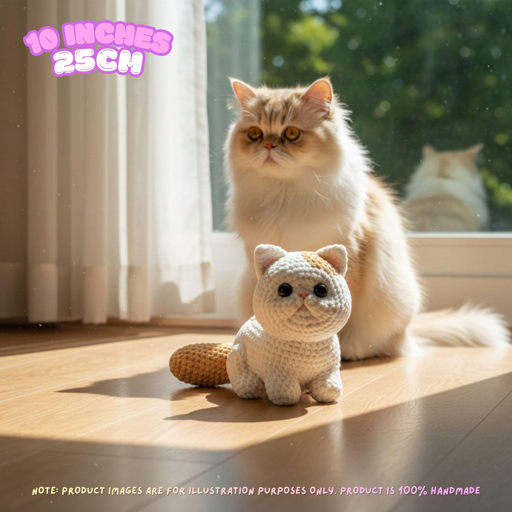 A small crocheted cat toy sits on a wooden floor in front of a fluffy cream-colored cat in a sunlit room. The image includes text overlays indicating the product size is 10 inches (25 cm) and a note that product images are for illustration purposes only.