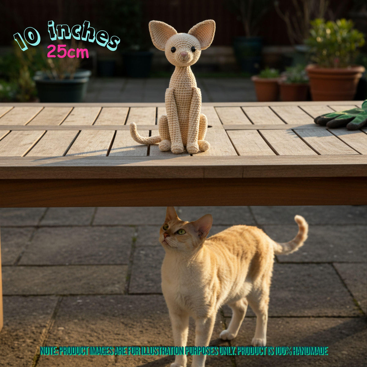 A crocheted cat-shaped plush toy sits on a wooden table in an outdoor patio setting, with a real cat standing underneath. Text in the top left reads '10 inches 25cm', and there is a note at the bottom about the product being handmade and for illustration purposes only.