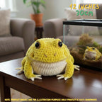 A yellow and cream crocheted frog plush toy sits on a wooden table in a living room setting, with a glass terrarium containing a real frog in the background. Text overlays indicate the size is 12 inches (30 cm) and note that the product image is for illustration purposes only.