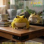 A yellow crocheted frog toy sits on a wooden table in a home setting, with its reflection visible in a glass terrarium behind it. Text in the top right reads '8 INCHES 20CM'.