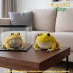 A yellow crocheted frog toy sits on a wooden table in a living room setting next to a small clear display case containing a real frog, with text overlays indicating the size of 6 inches (16 cm).