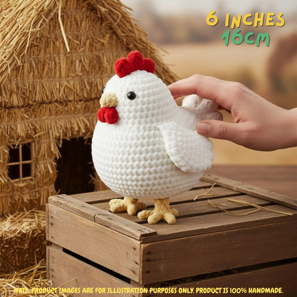 A white crocheted chicken toy with a red comb and yellow beak and feet, shown with a human hand touching it. The toy stands on a wooden crate in front of a straw henhouse, with text reading '6 INCHES 15CM' in the top right.