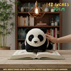 Hand gently touching top of a handmade giant panda bear crochet amigurumi plush doll lying on an open book on a wooden table in front of a bookshelf, with text reading 12 INCHES 30cm.