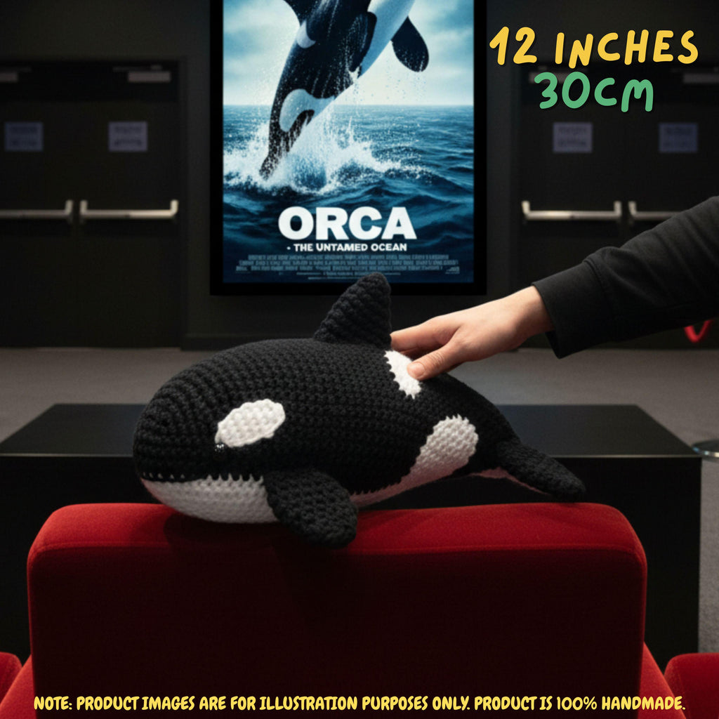 A crocheted orca plush toy resting on a red chair, with a human hand touching its back. A movie poster for 'Orca: The Untitled Ocean' is visible in the background, and text in the top right reads '12 INCHES 30CM'.