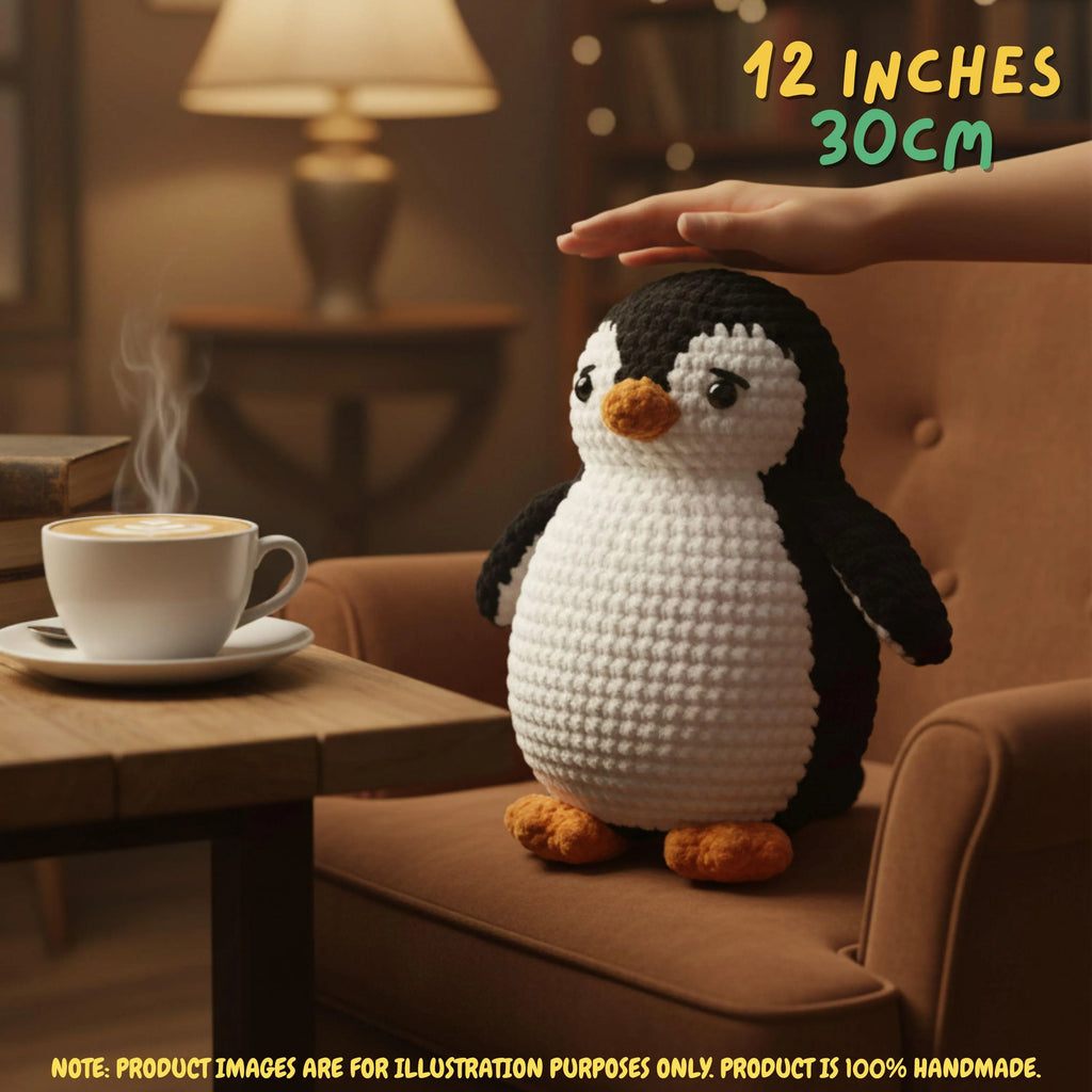 A crocheted penguin plush toy sits on a brown armchair in a warmly lit room, with a hand resting above its head. A coffee cup is on a table beside it, and text in the top right says '12 INCHES 30CM'.