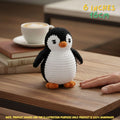 A small crocheted penguin toy sits on a wooden table next to a human hand, with a cup of coffee and books blurred in the background. Text in the top right says '6 INCHES 16CM' and a note at the bottom says the product is 100% handmade.