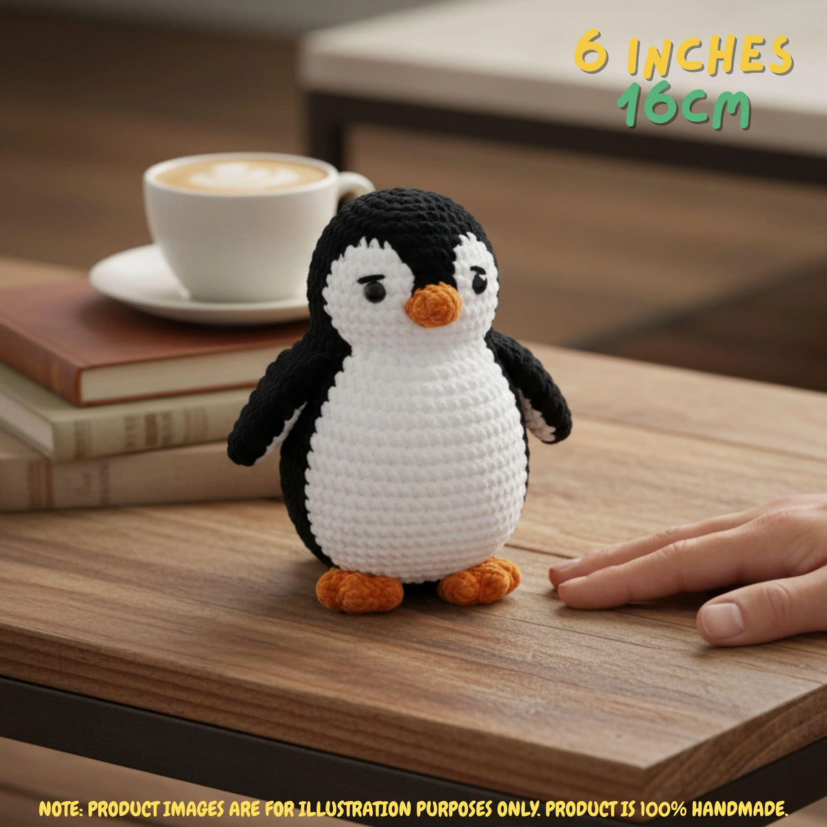 A small crocheted penguin toy sits on a wooden table next to a human hand, with a cup of coffee and books blurred in the background. Text in the top right says '6 INCHES 16CM' and a note at the bottom says the product is 100% handmade.