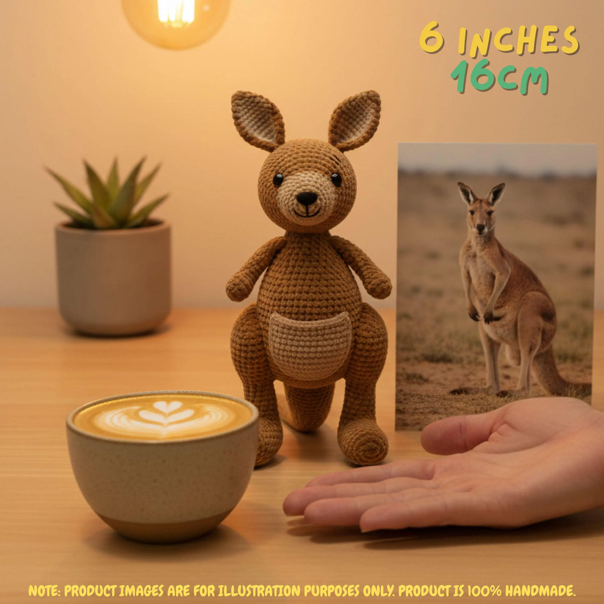 A crocheted kangaroo plush toy stands on a wooden table next to a cup of latte, a small potted plant, and a hand for scale, with a printed kangaroo photo in the background and text reading 6 inches / 16 cm.