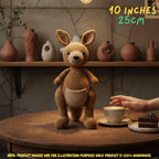 A crocheted kangaroo plush toy sits on a wooden table in a cozy indoor setting, with a hand reaching toward a cup nearby. The image includes text overlays stating '10 INCHES 25CM' and a note that the product images are for illustration purposes only and the product is 100% handmade.