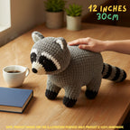 A crocheted raccoon toy is shown on a wooden table with a human hand resting on its back, a white coffee cup, a blue notebook, and potted plants in the background. Text in the top right reads '12 INCHES 30CM'.