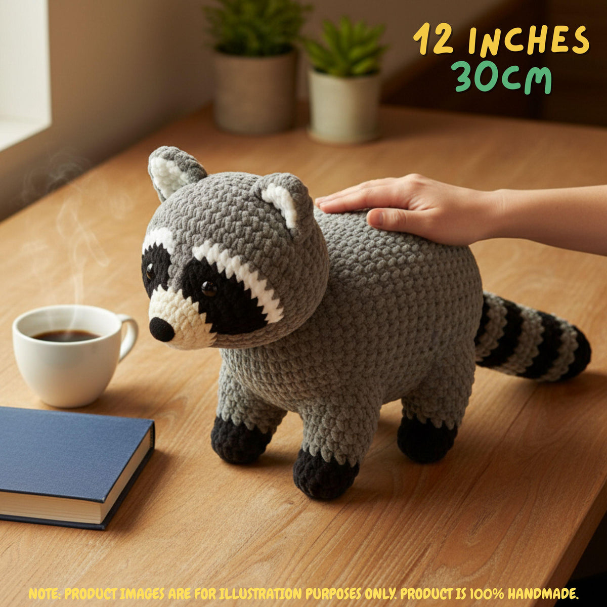 A crocheted raccoon toy is shown on a wooden table with a human hand resting on its back, a white coffee cup, a blue notebook, and potted plants in the background. Text in the top right reads '12 INCHES 30CM'.