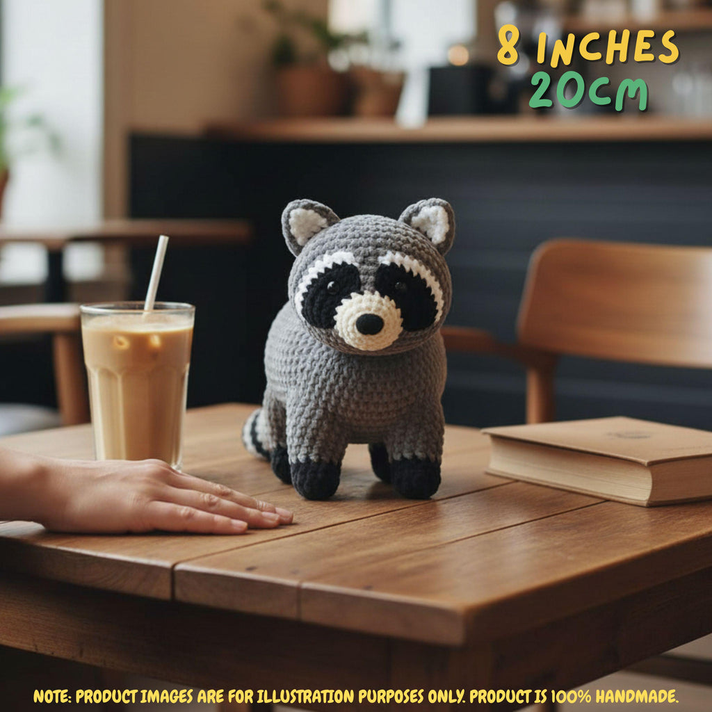A crocheted raccoon toy sits on a wooden table in a cafe-like setting, with a hand near it, a drink, and a book around it. Text in the top right reads '8 INCHES 20CM' and a note at the bottom says the product image is for illustration purposes only and the product is 100% handmade.