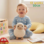 A smiling toddler in a light blue cloud-print pajama set sits on a play mat holding a crocheted sheep toy. The scene includes a wooden alphabet box, a yellow crescent pillow, and text overlays reading '12 INCHES 30CM' and a product note at the bottom.