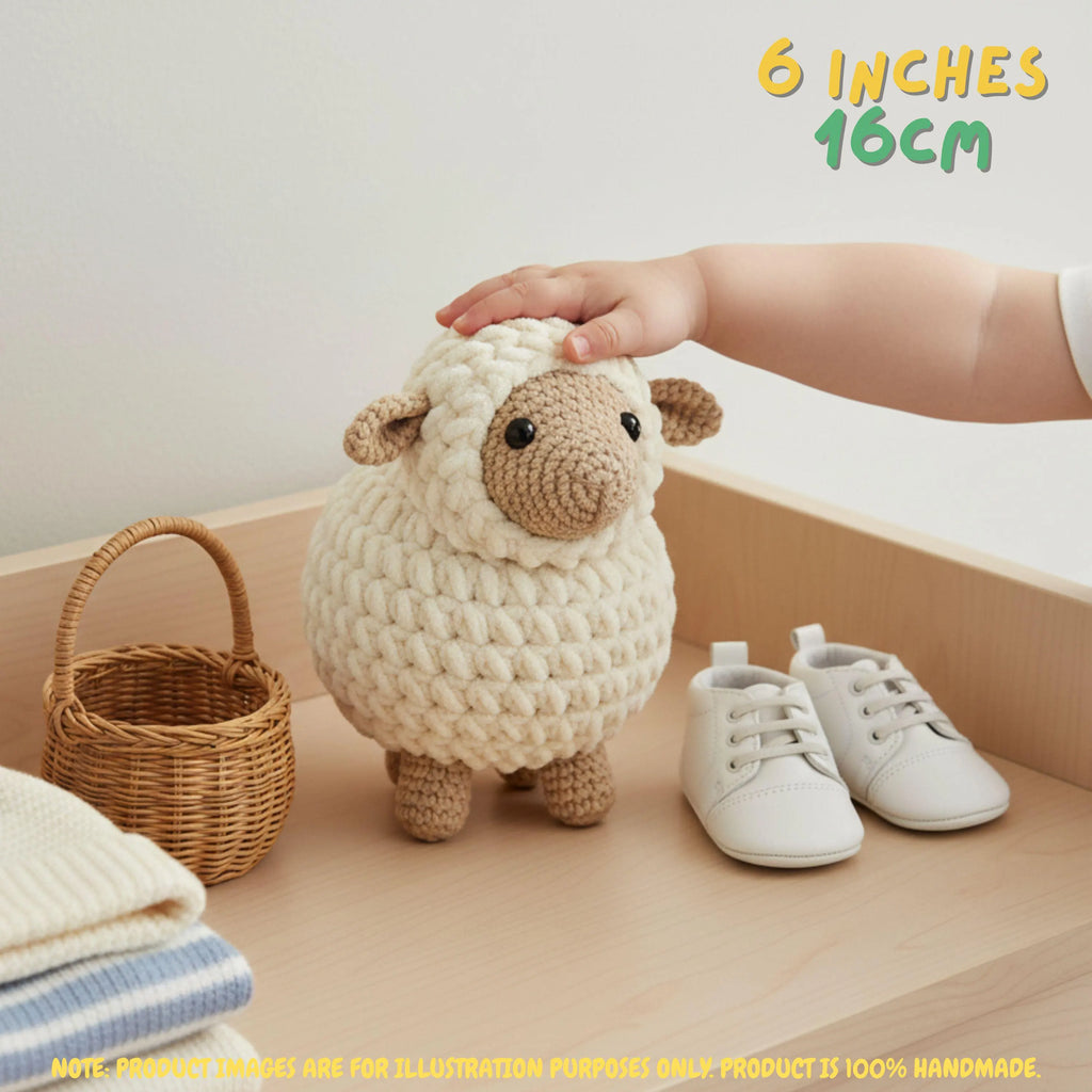 A crocheted sheep toy with a cream body and tan face is displayed on a wooden shelf next to a small wicker basket, white shoes, and folded blankets. A human hand is resting on the sheep's head, and text in the top right reads '6 INCHES 16CM'.