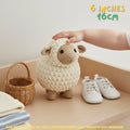 A crocheted sheep toy with a cream body and tan face is displayed on a wooden shelf next to a small wicker basket, white shoes, and folded blankets. A human hand is resting on the sheep's head, and text in the top right reads '6 INCHES 16CM'.