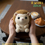 A crocheted hedgehog plush toy being held in two hands over a wooden table, with a coffee cup and croissant in the background. Text in the top right reads '8 INCHES 20cm' and a note at the bottom says product images are for illustration purposes only.