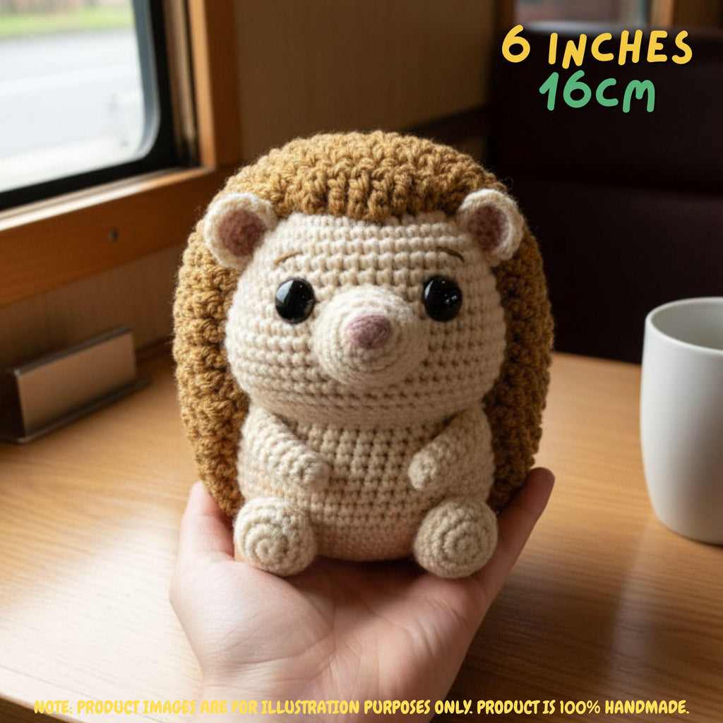 A crocheted hedgehog toy being held in a person's hand on a wooden table, with a window and white mug in the background. Text in the top right reads '6 INCHES 16CM' and there is a note at the bottom stating product images are for illustration purposes only.