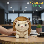 A crocheted hedgehog plush toy being held by a person's hand on a wooden table, with a blurred cafe interior in the background. Text overlays read '10 INCHES 25CM' and 'NOTE.