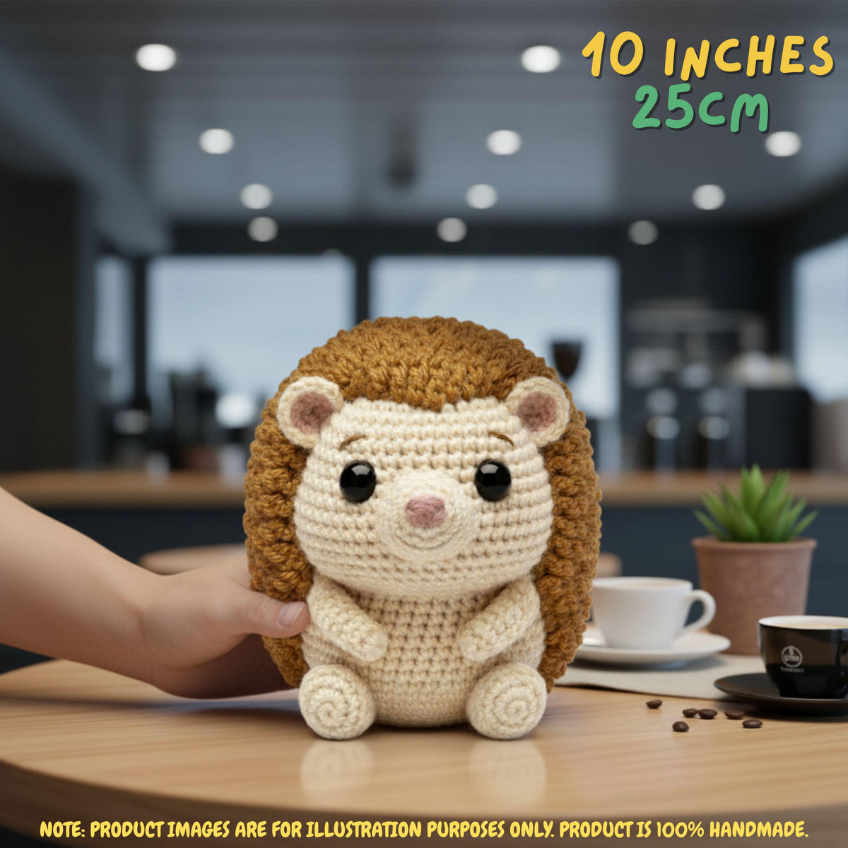 A crocheted hedgehog plush toy being held by a person's hand on a wooden table, with a blurred cafe interior in the background. Text overlays read '10 INCHES 25CM' and 'NOTE.