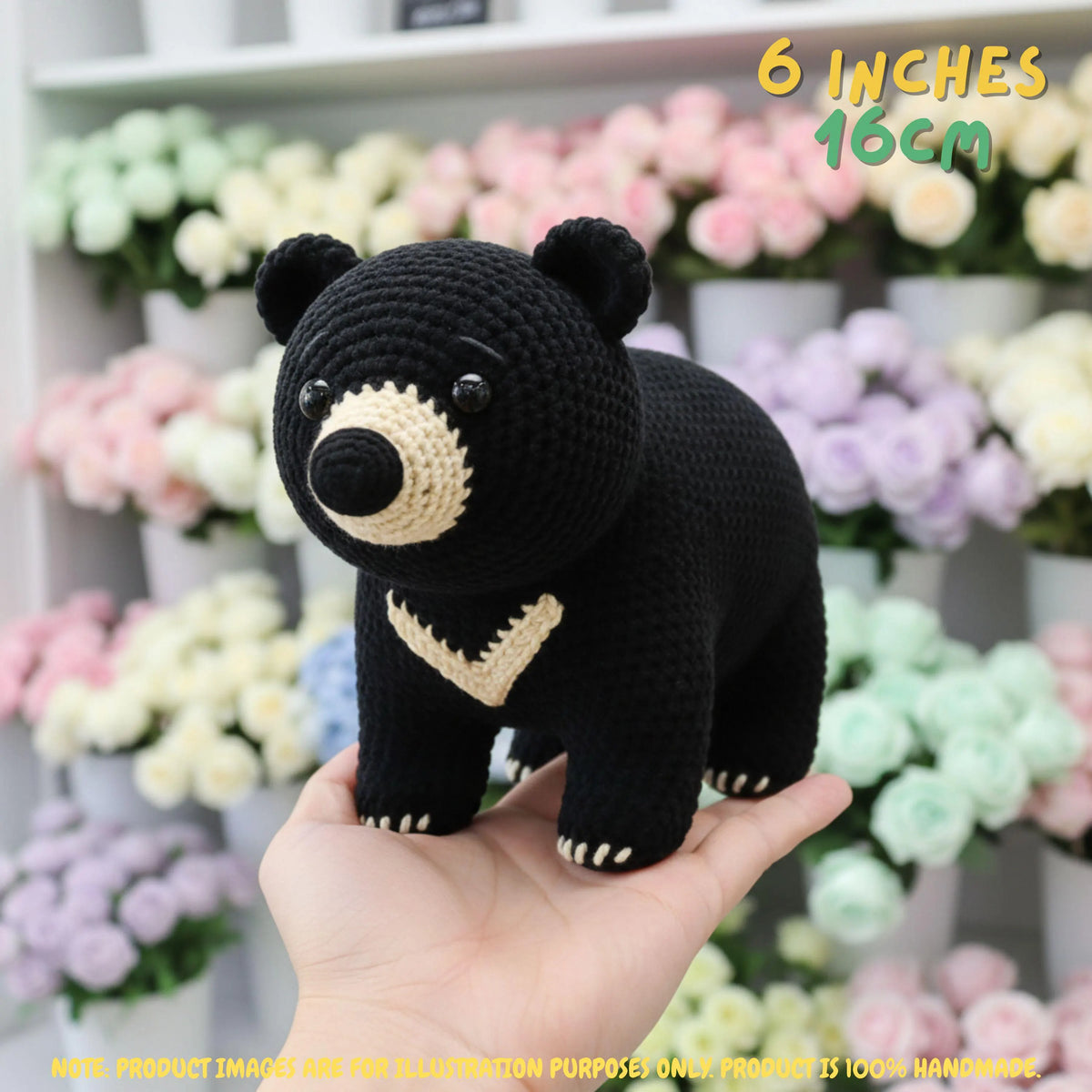 A crocheted black bear plush toy with beige accents is held in a person's hand, with a blurred background of pastel flowers. Text in the top right reads '6 INCHES 16CM'.