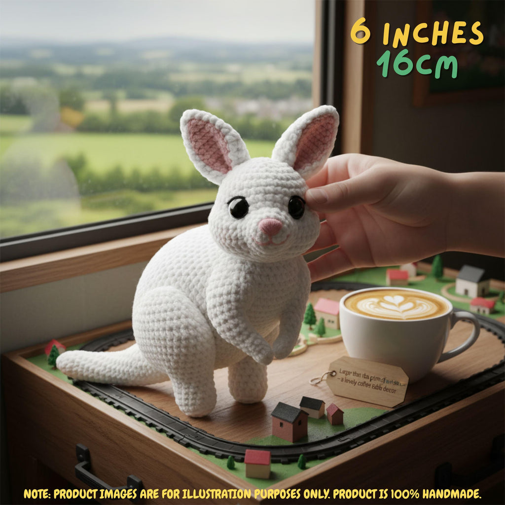 A white crocheted kangaroo toy is shown on a miniature train track beside a cup of coffee, with a human hand touching the toy. Text in the corner says 6 inches 16cm.