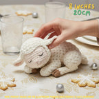 Handmade cream-colored crochet baby lamb amigurumi doll in a sleeping pose held by a hand on a table with holiday cookies, featuring "8 INCHES / 20CM" text overlay.