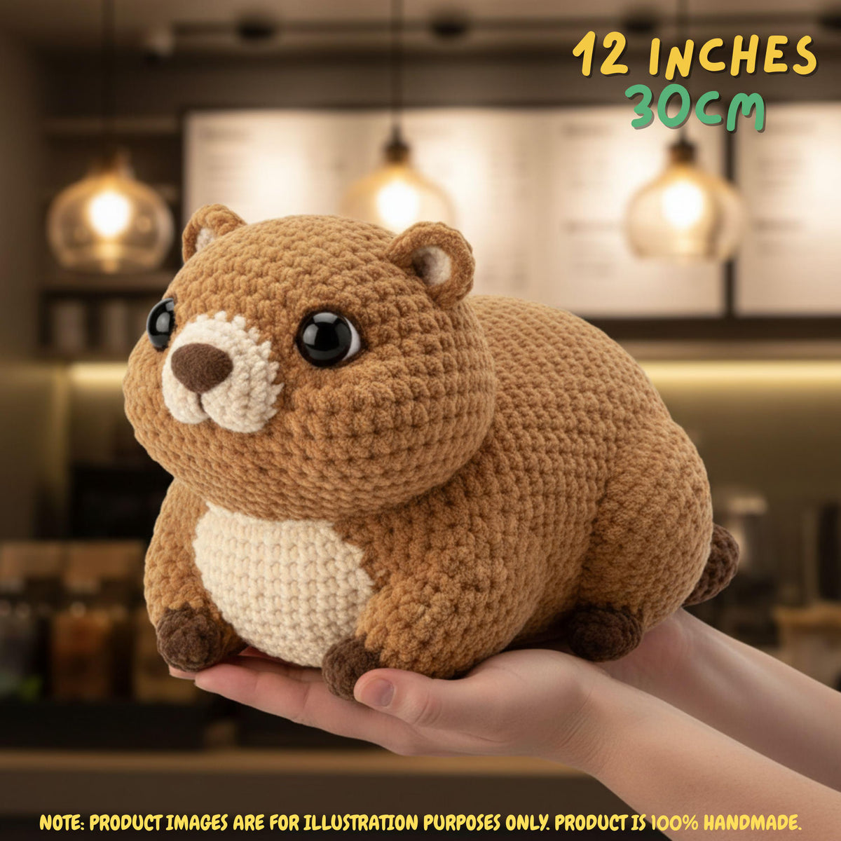 A crocheted brown animal plush toy with a cream belly and black eyes, being held in a person's hands. The background is a softly blurred cafe interior with hanging lights.